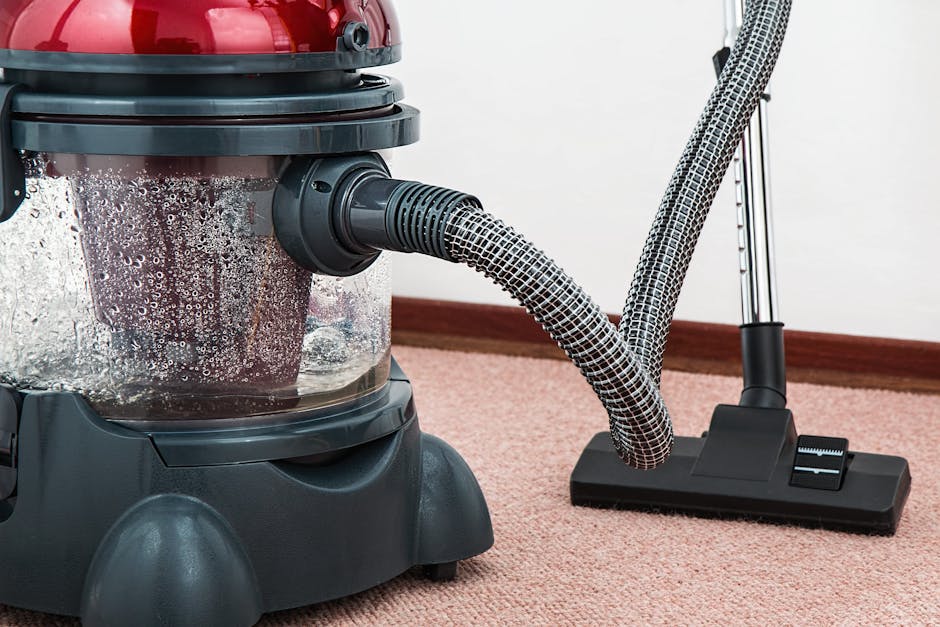 A professional cleaner dressed in full white protective suit, mask, and blue gloves is performing a deep cleaning process on a beige carpet inside a modern living room. The cleaner is using a speciali